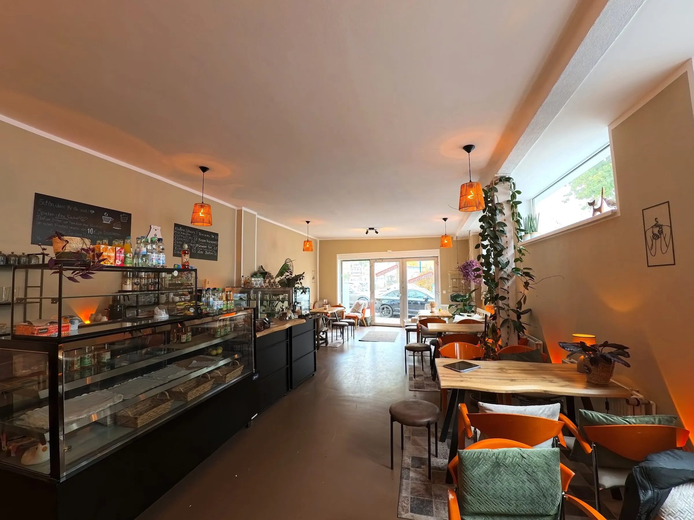 Der lange Café-Raum mit Kuchenvitrine, Holztischen, orangen Stühlen und Pflanzen